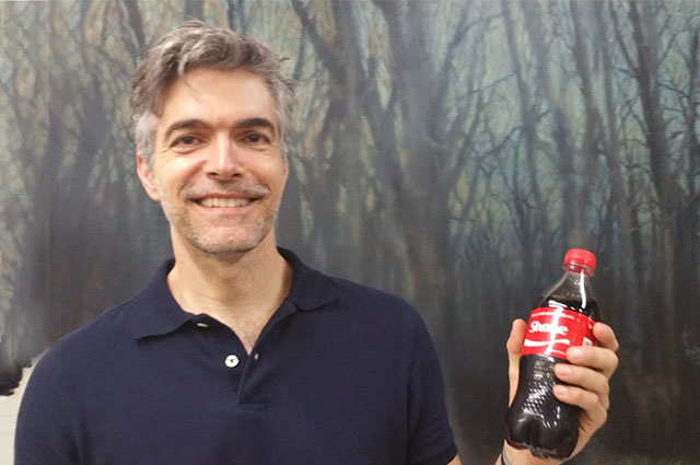 Shane stands with a custom Coke bottle and a smile in front of one of his paintings.