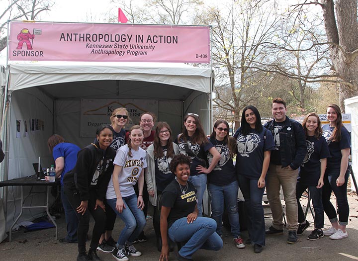 12 KSU students posing in front of "Anthropology In Action" sign and activity tent at Atlanta Science Festival Expo