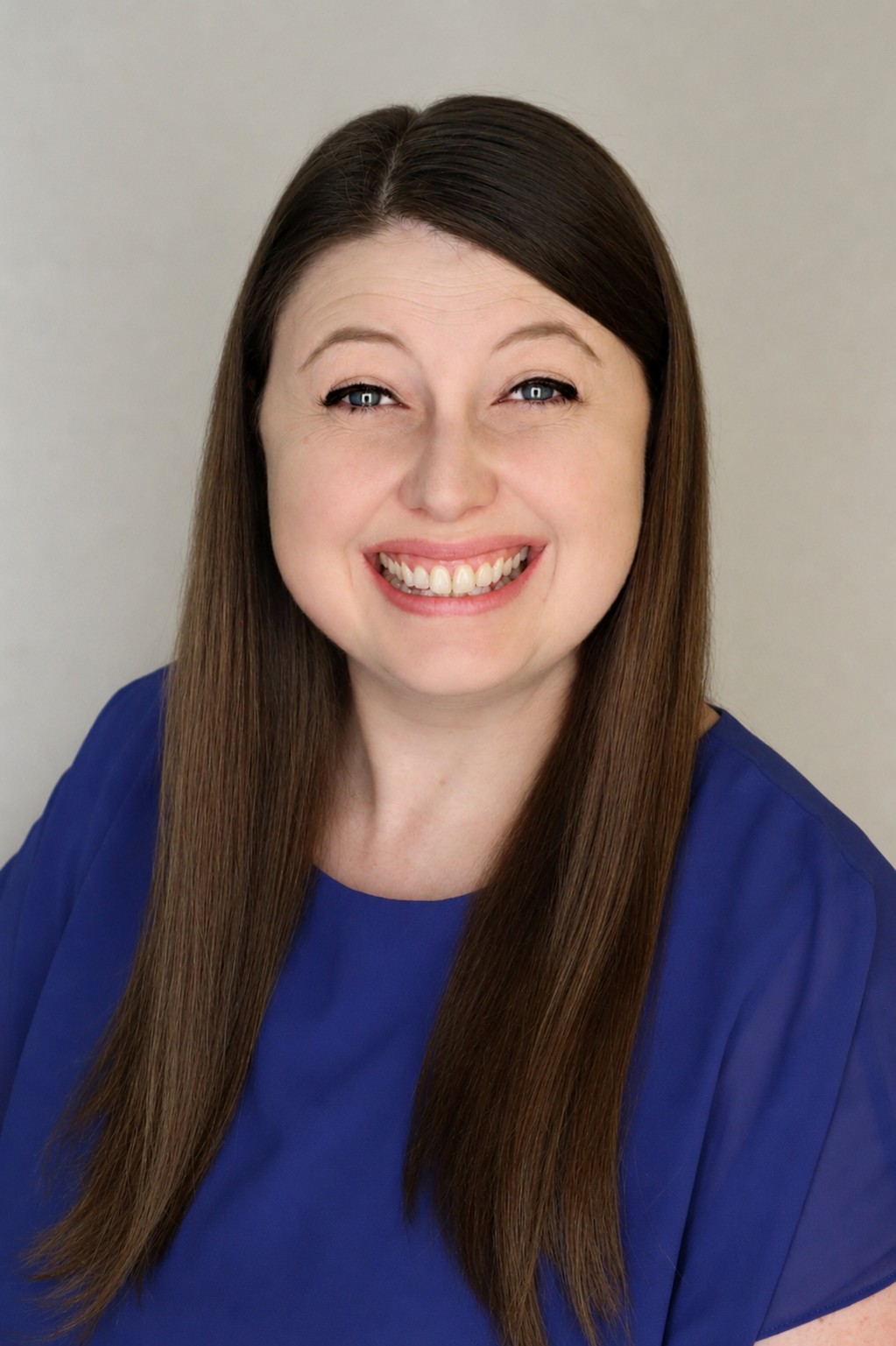 Professional headshot of a smiling woman with long, straight brown hair wearing a blue blouse, posed against a light neutral background.