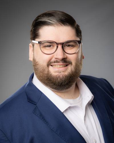 Headshot of a White male with dark hair and a beard, wearing glasses and a blue suit coat