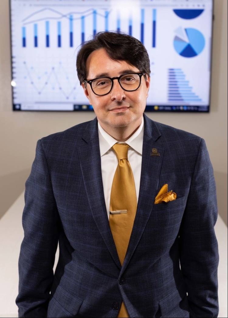 Man with dark hair and glasses in blue blazer and gold tie